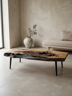 A rustic live-edge wooden coffee table with black metal legs, styled in a minimalist, warm-toned modern living room next to a concrete wall and a neutral sofa.
