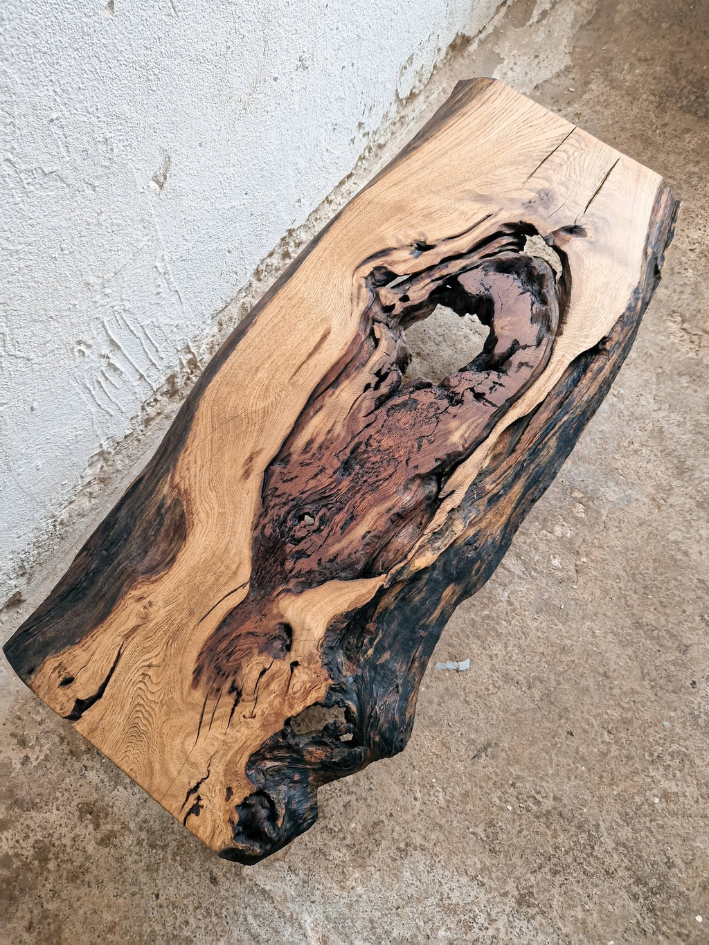 A rustic live-edge wooden slab with natural bark and rich wood grain patterns, photographed on a concrete floor against a textured wall.
