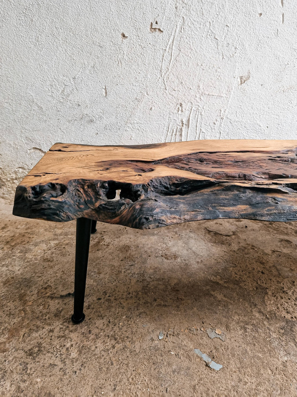 A rustic live-edge wooden table with a natural bark-look surface and a black metal leg, photographed indoors against a textured off-white wall and concrete floor.