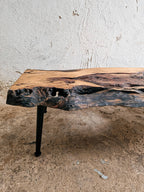 A rustic live-edge wooden table with a natural bark-look surface and a black metal leg, photographed indoors against a textured off-white wall and concrete floor.