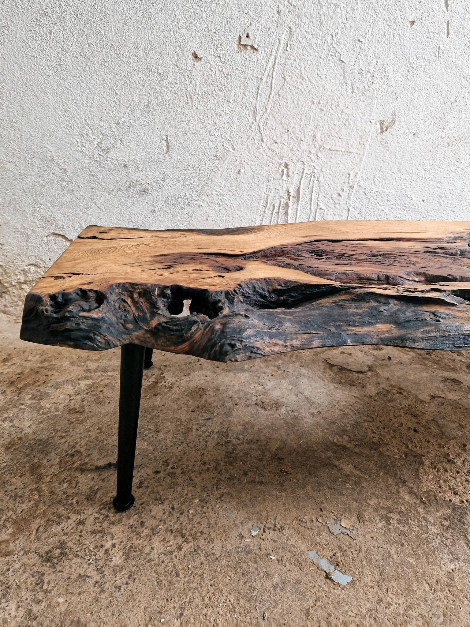 A rustic live-edge wooden table with a natural bark-look surface and a black metal leg, photographed indoors against a textured off-white wall and concrete floor.