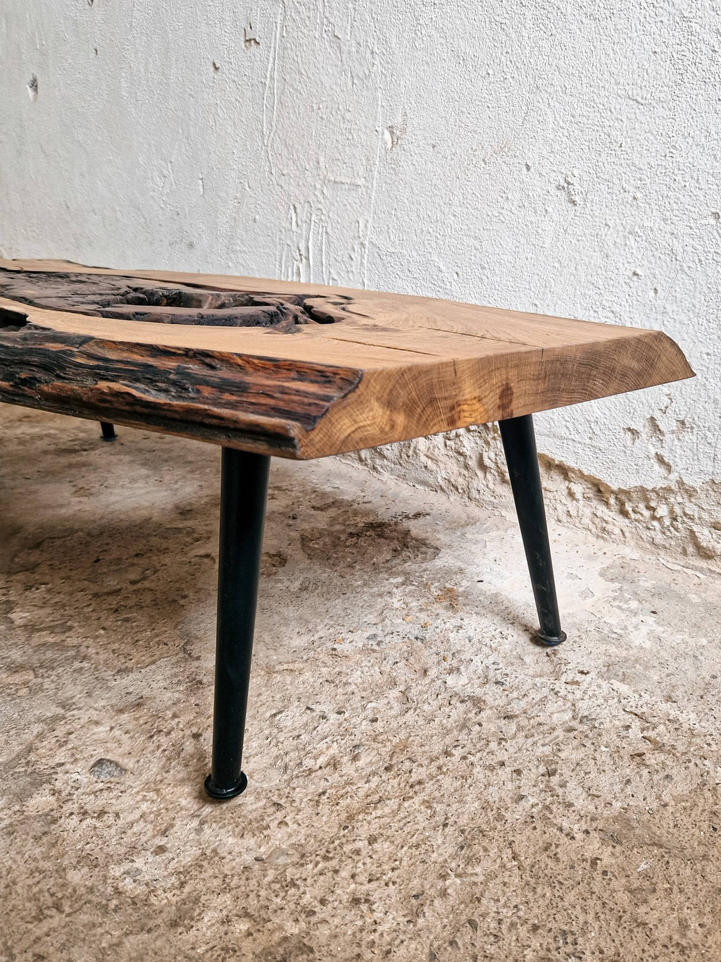 A rustic live-edge wooden coffee table with a dark natural wood top and black metal legs, shown in a textured room setting.