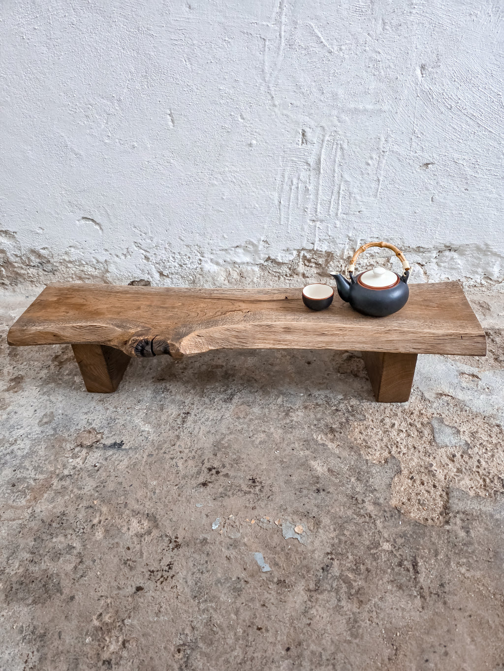 Wooden bench with a teapot and cup on a concrete floor against a white wall.